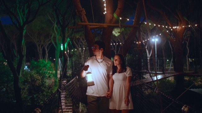 Una coppia cammina su un ponte sospeso illuminato da luci, lui tiene una lanterna.