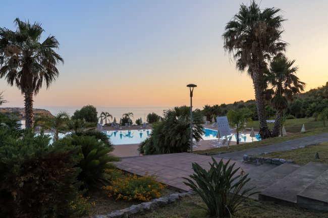 Piscina al tramonto circondata da palme e vista mare.