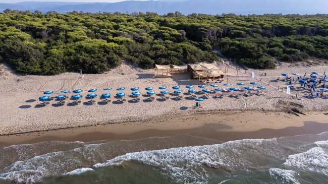 Vista aerea della spiaggia con ombrelloni blu e un chiosco, circondata da vegetazione.