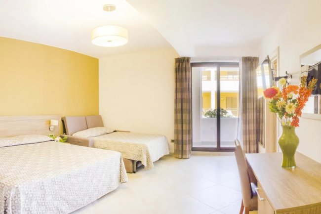 Camera con letto matrimoniale e singolo. Finestra aperta, tavolo con vaso di fiori colorati.