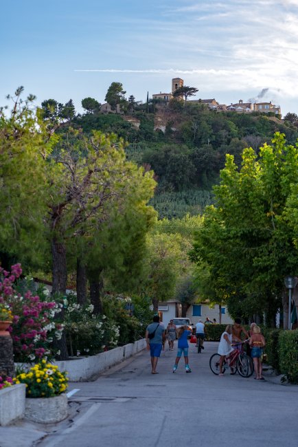 Strada del campeggio con persone e biciclette, vista di un paese su una collina sullo sfondo.