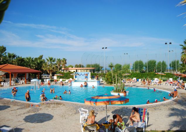 Piscina affollata circondata da lettini, ombrelloni e palme. Cielo sereno.