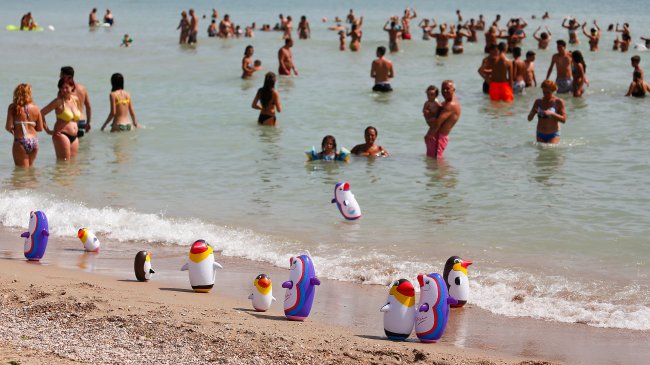 Spiaggia affollata con persone in acqua e giochi a forma di pinguino sulla sabbia.
