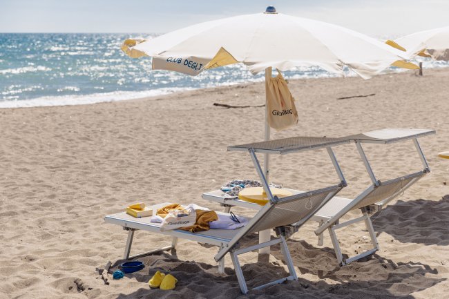 Due lettini con ombrellone bianco su una spiaggia sabbiosa, accessori sparsi.