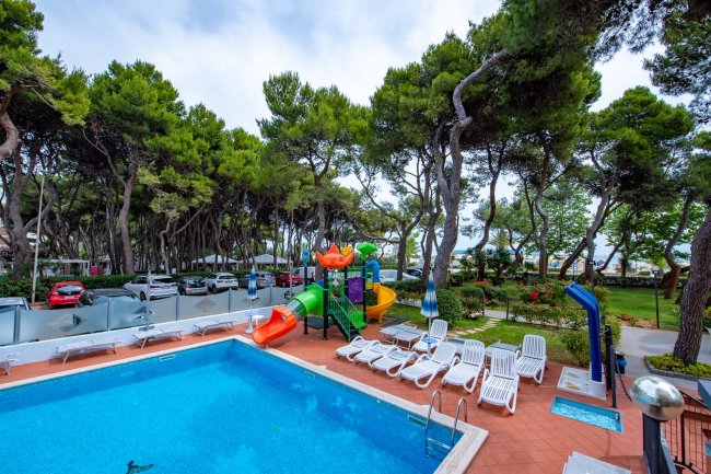 Piscina all'aperto con scivoli colorati e lettini sotto alberi.