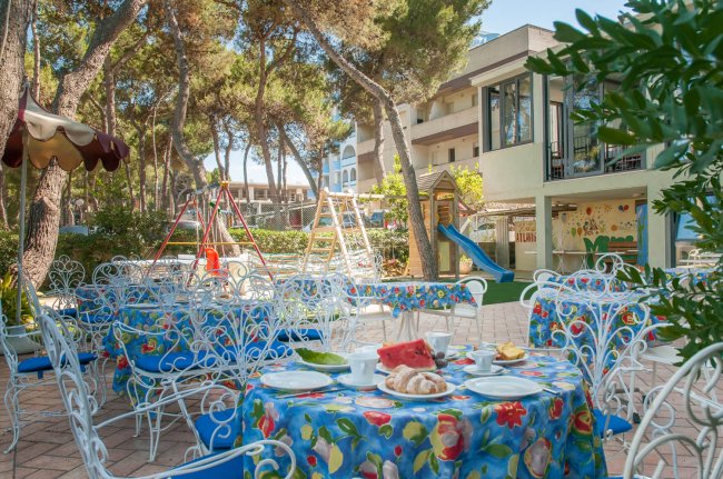 Tavoli colorati in un giardino con parco giochi tra gli alberi.