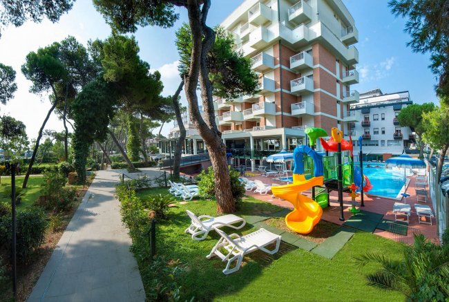 Un hotel con parco giochi colorato, piscina e sedie a sdraio all'aperto, circondato da alberi.