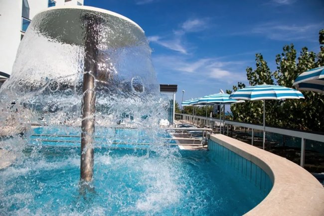 Una fontana a cascata in una piscina, con cielo blu e ombrelloni a righe sullo sfondo.