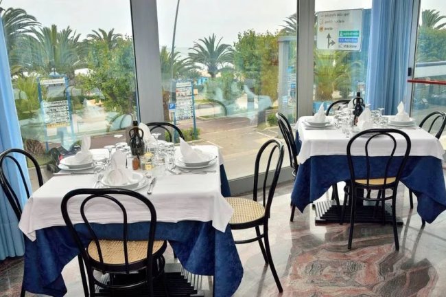 Interno di un ristorante con tavoli apparecchiati e grandi finestre che mostrano palme esterne.