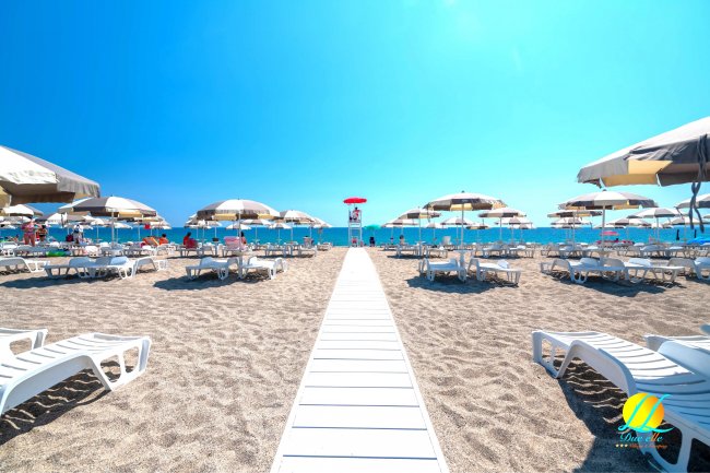 Passerella bianca su sabbia, circondata da lettini e ombrelloni, verso il mare.