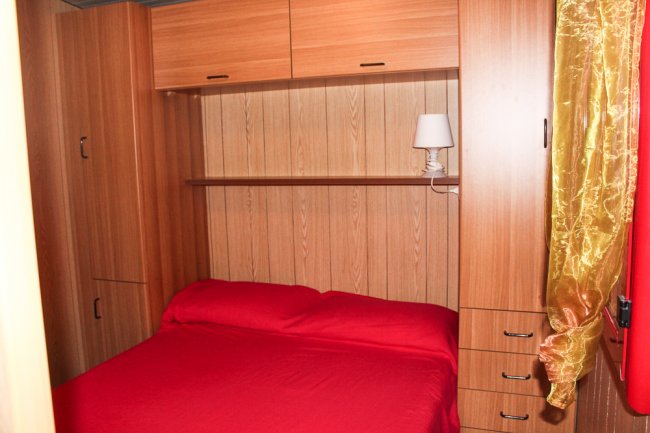Camera da letto con mobili in legno, letto matrimoniale con coperta rossa e lampada su mensola.