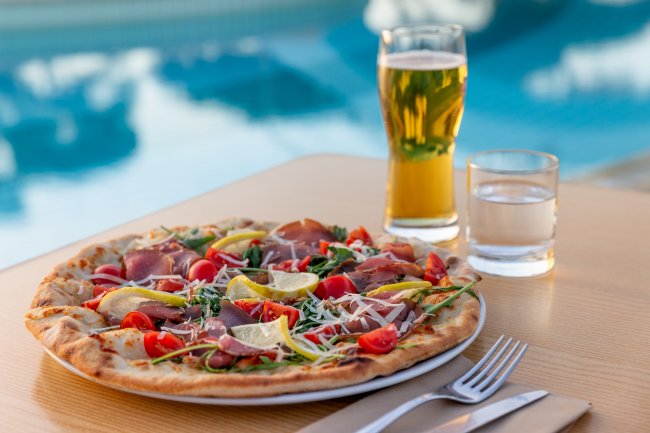 Pizza con verdure e prosciutto su un tavolo, birra e acqua, vista piscina.