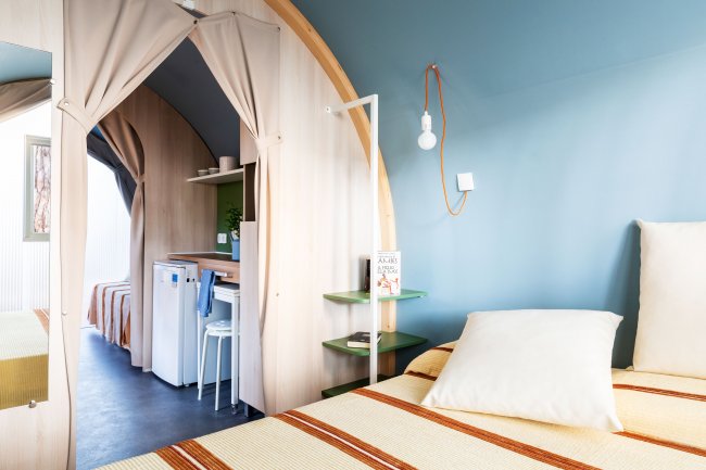 Interno moderno di una cabina con letto, mini-frigo e scaffali, decorato con toni chiari.
