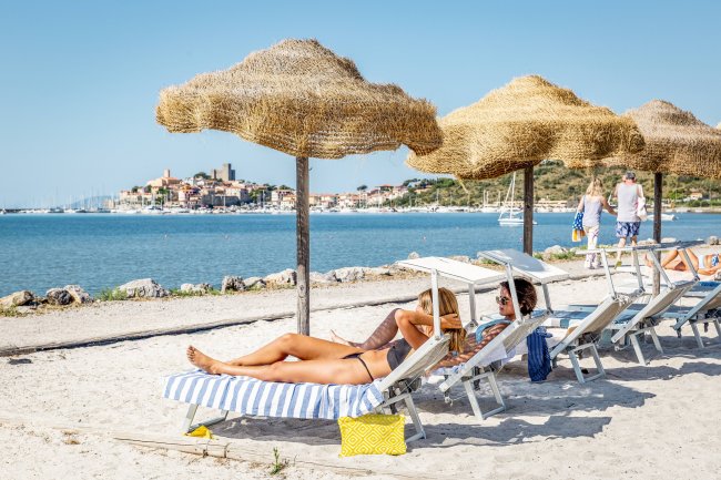 Persone su lettini sotto ombrelloni di paglia, vista su mare e borgo in lontananza.