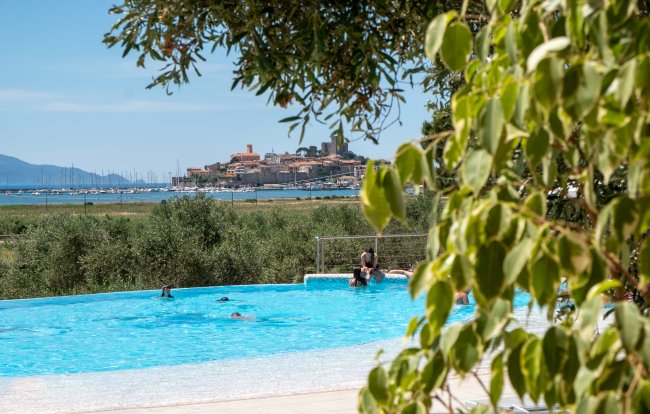 Piscina all'aperto con vista su un villaggio costiero in lontananza, e foglie verdi in primo piano.