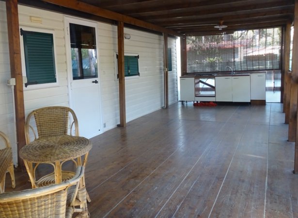 Veranda in legno con sedie e tavolo in rattan, cucina esterna sullo sfondo.
