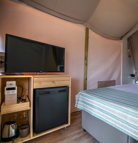 Interno di una tenda glamping con TV, mini-frigo e letto coperto da trapunta a righe blu.