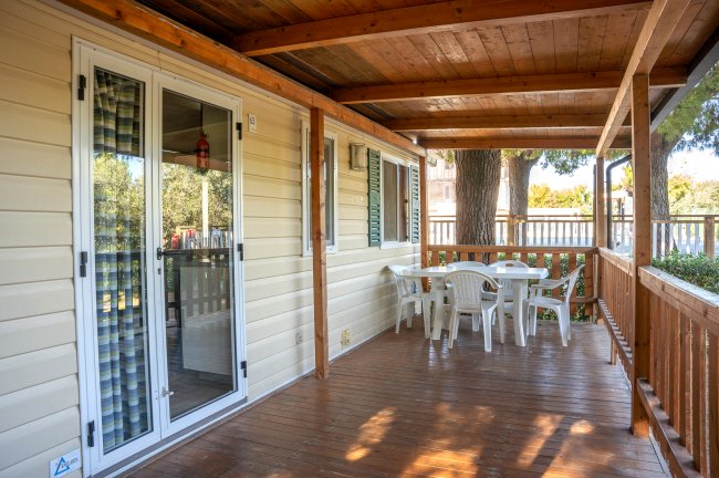 Veranda con tetto in legno, tavolo e sedie bianche, vista su giardino e alberi.