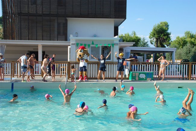 Piscina con bambini che nuotano e adulti sul bordo. Una persona in costume da mascotte sullo sfondo.