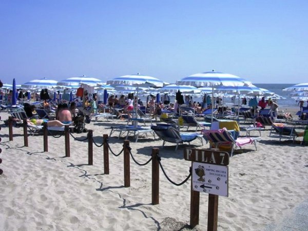 Spiaggia affollata con lettini, ombrelloni bianchi e persone che prendono il sole.