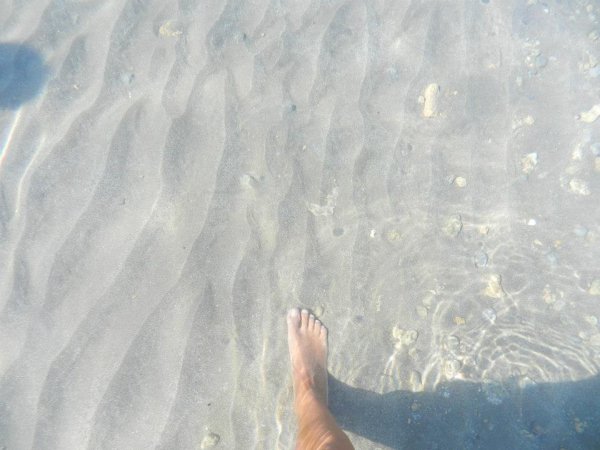 Un piede nell'acqua trasparente sopra sabbia ondulata.