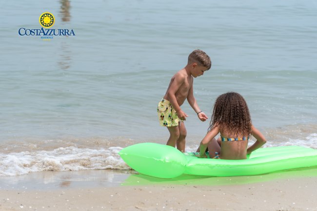 Due bambini giocano sulla spiaggia, uno seduto su un materassino verde in acqua.