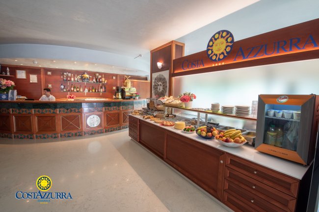 Lobby luminosa con un bancone bar e un tavolo con frutta fresca e stoviglie.