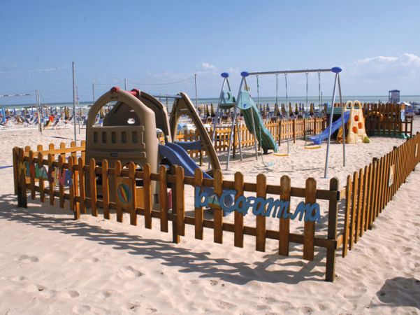 Area giochi per bambini su spiaggia sabbiosa, con altalene e scivoli circondati da una staccionata.
