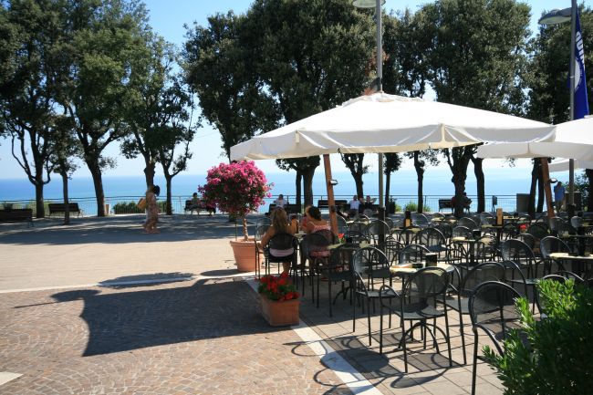 Tavolini all'aperto sotto ombrelloni bianchi, vista mare e alberi in lontananza.