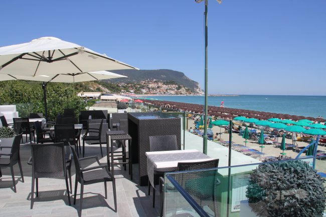 Terrazza con sedie e tavoli sotto ombrelloni, vista mare e spiaggia affollata.