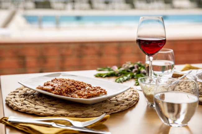 Tavola apparecchiata con piatto di cibo e bicchiere di vino rosso vicino a una piscina.