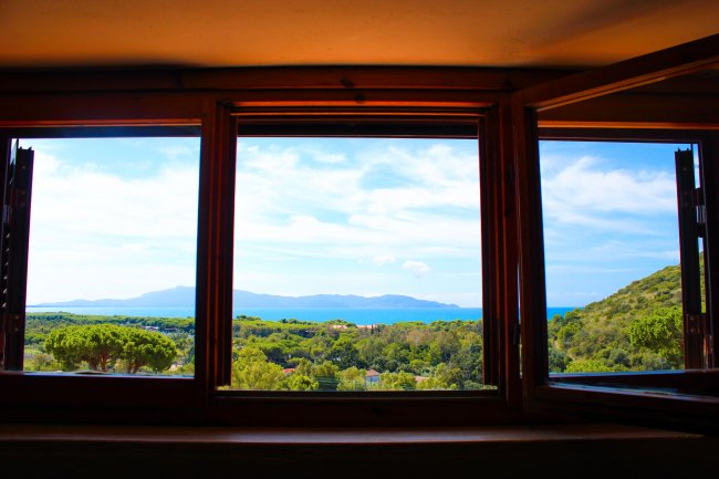 Finestra aperta con vista su colline verdi, mare e cielo azzurro con nuvole.
