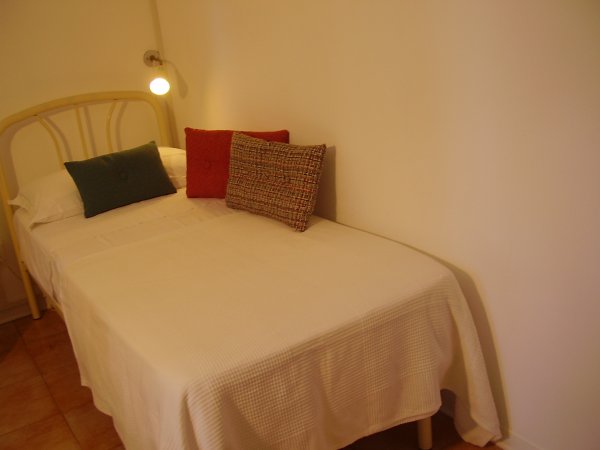 Letto singolo con tre cuscini colorati su copriletto bianco, sotto una luce.