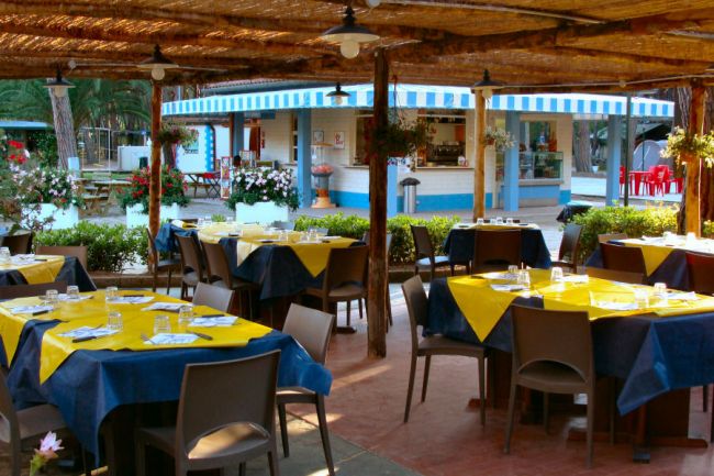 Area ristorante all'aperto con tavoli apparecchiati in giallo e blu, con chiosco sullo sfondo.