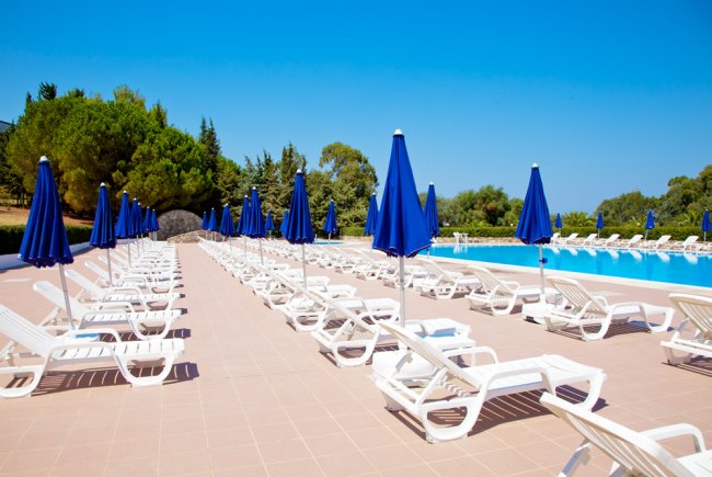 Piscina con lettini bianchi e ombrelloni blu, cielo sereno.