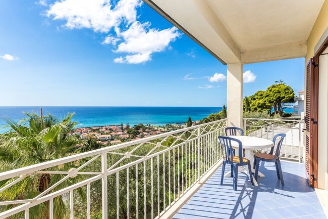 Balcone con tavolino e due sedie affacciato sul mare, vista panoramica su palme e tetti.