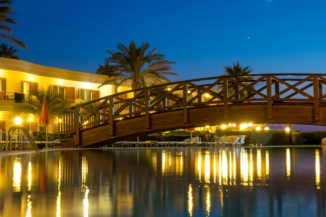 Ponte in legno illuminato sopra una piscina di notte, con palme sullo sfondo.
