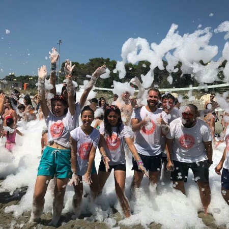 Persone sorridenti in una festa con schiuma all'aperto, tutti bagnati e festosi.