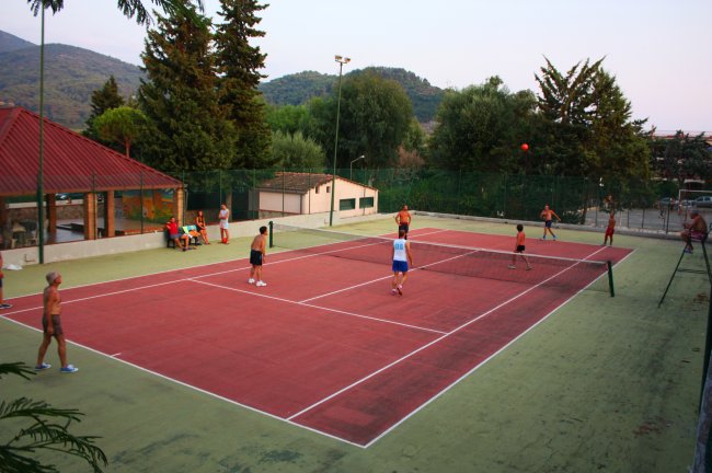 Persone giocano a tennis su un campo all'aperto circondato da alberi.