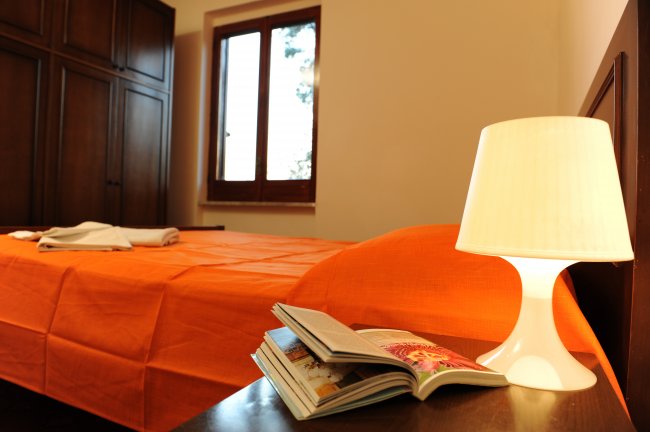 Camera con letto matrimoniale, copriletto arancione, lampada da tavolo e libro aperto.