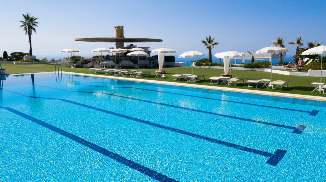 Una piscina con ombrelloni bianchi, prato verde e vista mare sullo sfondo.