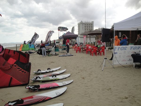 Spiaggia con tavole da surf, sedie rosse e striscioni esposti in una giornata nuvolosa.