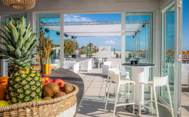 Sala con tavoli bianchi e panche, vista esterna mare. Frutta fresca in primo piano.