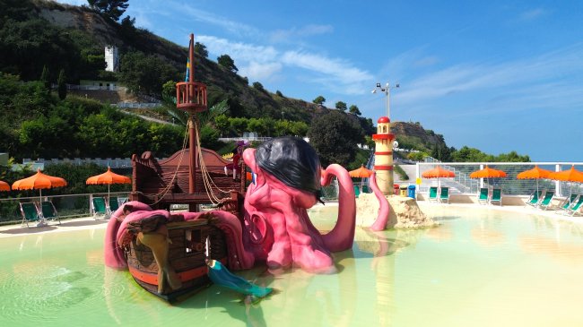 Piscina con struttura a forma di polpo gigante rosa vicino a un faro e ombrelloni arancioni.