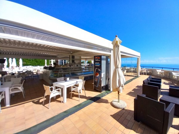 Un bar all'aperto con tavoli bianchi e ombrelloni chiusi su una terrazza soleggiata.