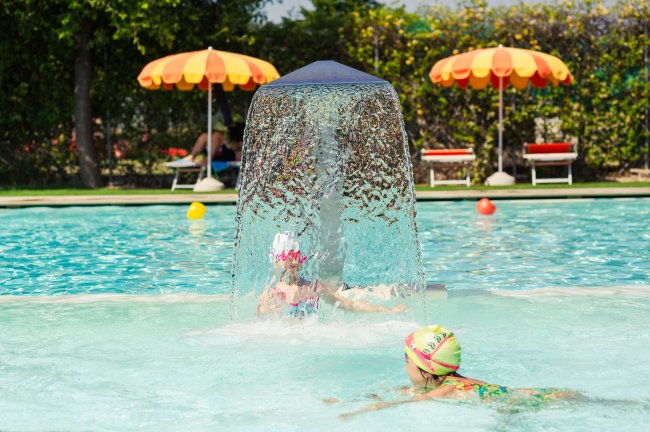 Due bambine giocano in piscina sotto una fontana d'acqua. Ombrelloni sullo sfondo.