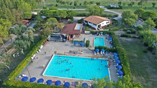 Piscina con persone e ombrelloni blu, circondata da alberi e edifici.